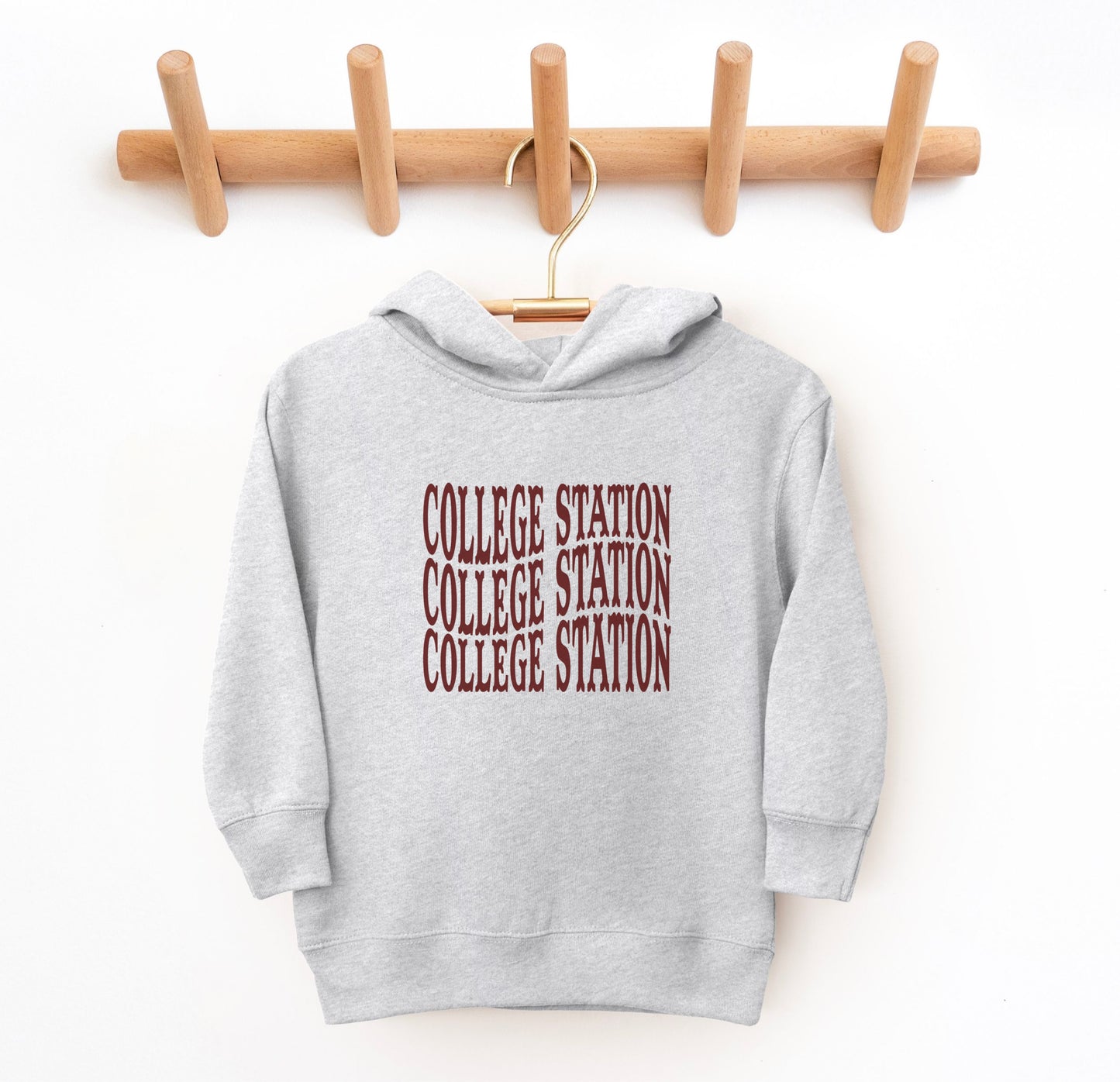 The Heather Grey Toddler Unisex College Station Western Hooded Sweatshirt lays flat on a white background. The ﻿College Station Western﻿ graphic is in bold Maroon in a Western style.