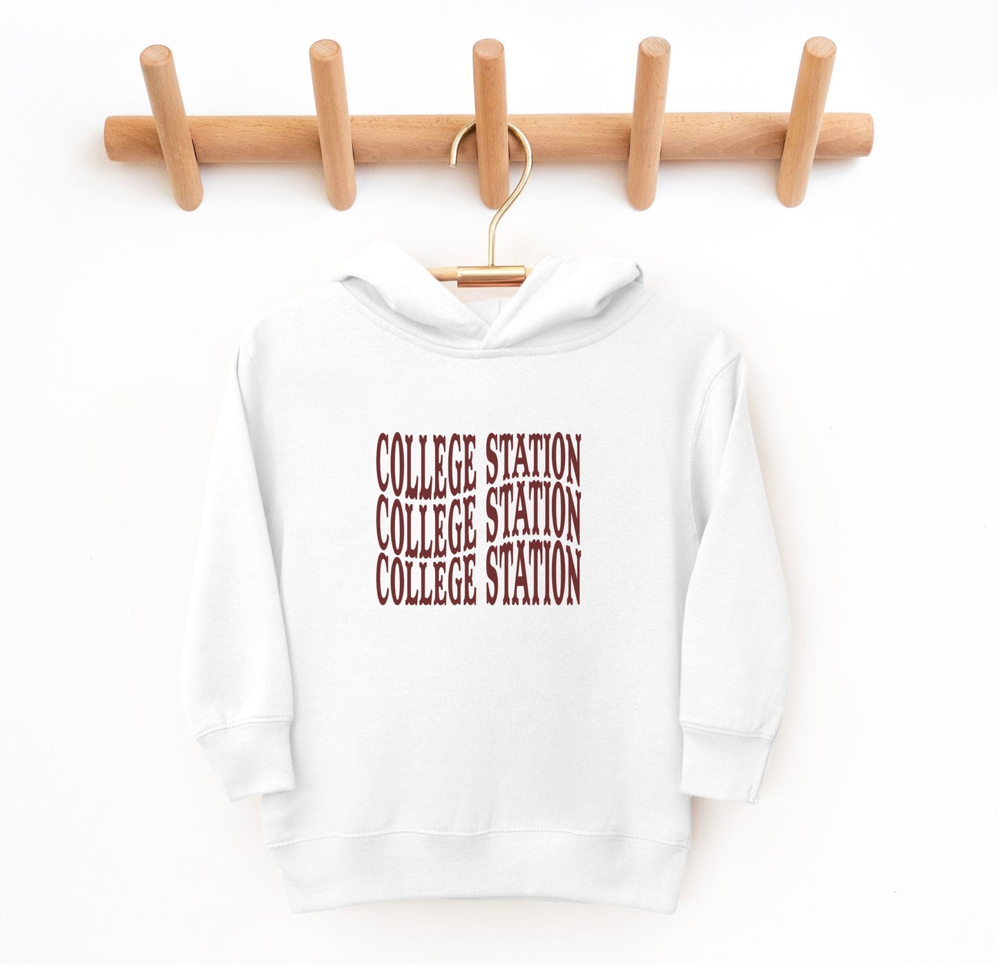 The White Toddler Unisex College Station Western Hooded Sweatshirt lays flat on a white background. The ﻿College Station Western﻿ graphic is in bold Maroon in a Western style.