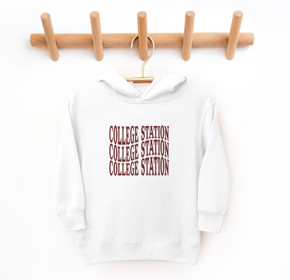 The White Toddler Unisex College Station Western Hooded Sweatshirt lays flat on a white background. The ﻿College Station Western﻿ graphic is in bold Maroon in a Western style.