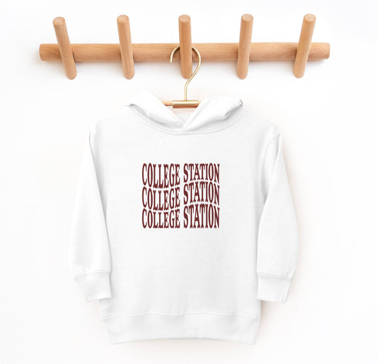 The White Toddler Unisex College Station Western Hooded Sweatshirt lays flat on a white background. The ﻿College Station Western﻿ graphic is in bold Maroon in a Western style.