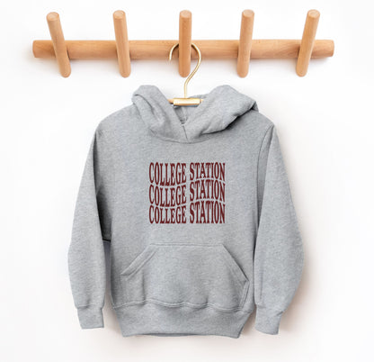 The Heather Youth Unisex College Station Western Hooded Sweatshirt lays flat on a white background. The ﻿College Station Western﻿ graphic is in bold Maroon in a Western style.