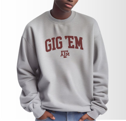 A model wears the Sport Grey Adult Unisex Texas A&M Gig 'Em Collegiate Crewneck Sweatshirt.  The ﻿Texas A&M Gig 'Em Collegiate﻿ graphic is in bold Maroon in a Collegiate style.