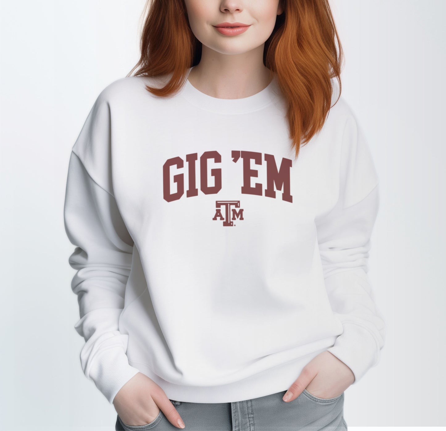 A model wears the White Adult Unisex Texas A&M Gig 'Em Collegiate Crewneck Sweatshirt.  The ﻿Texas A&M Gig 'Em Collegiate﻿ graphic is in bold Maroon in a Collegiate style.