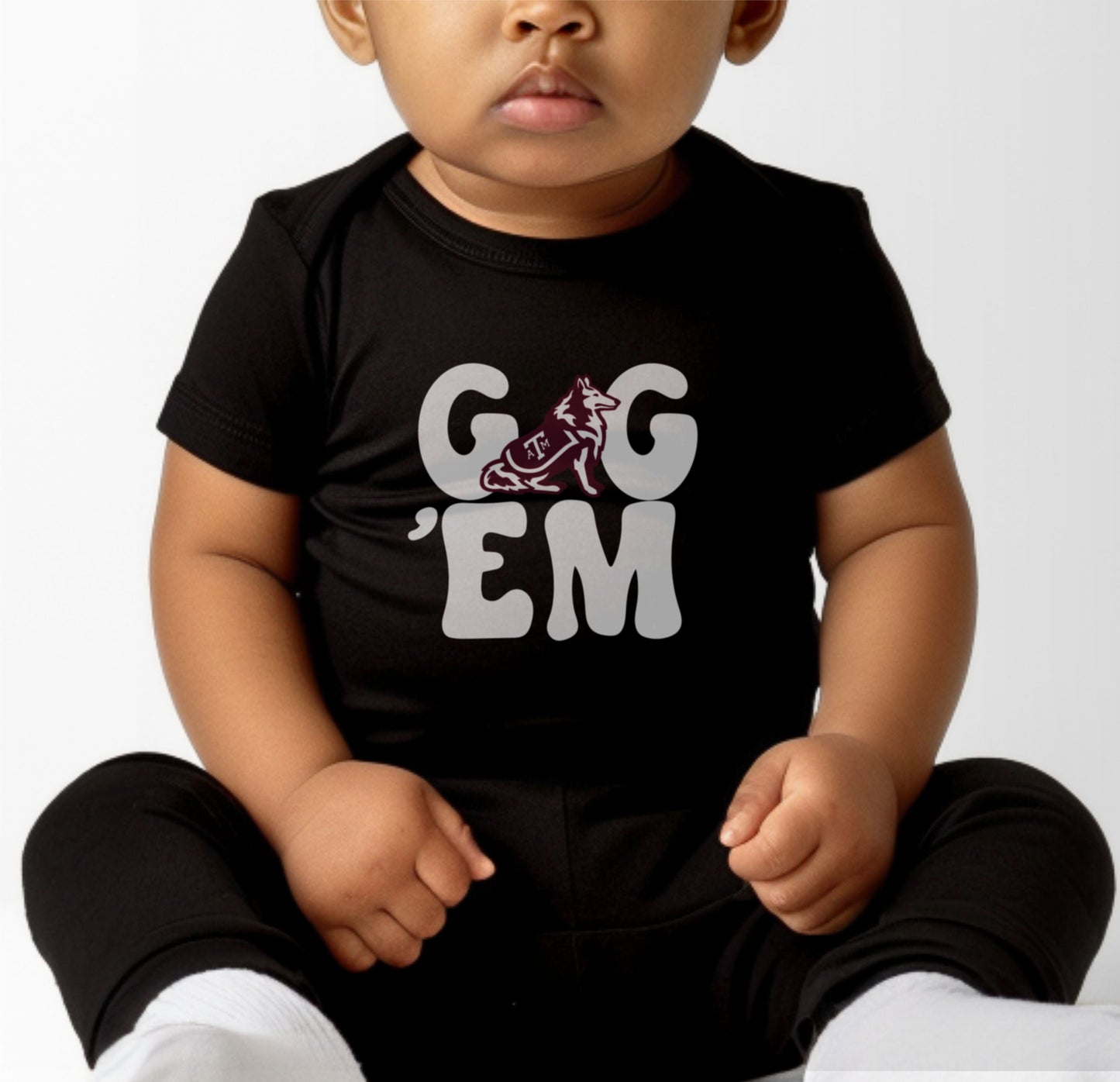 A model wears the Heather Grey Infant Unisex Texas A&M Gig 'Em Retro Reveille Bodysuit.  The ﻿Texas A&M Gig 'Em Retro Reveille﻿ graphic is in bold Maroon in a Groovy Vintage style.