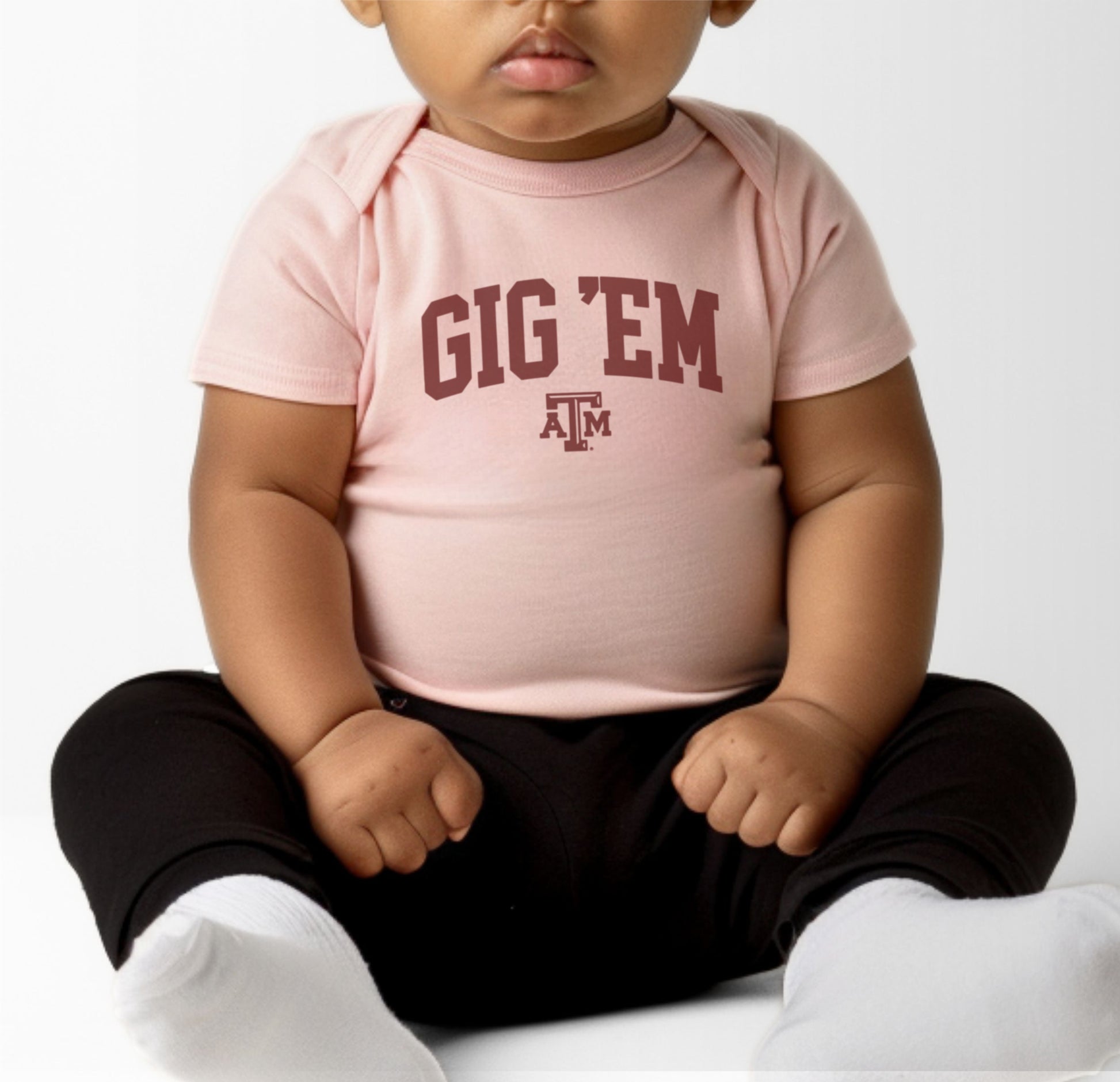 A model wears the Heather Grey Infant Unisex Texas A&M Gig 'Em Collegiate Bodysuit.  The ﻿Texas A&M Gig 'Em Collegiate﻿ graphic is in bold Maroon in a Collegiate style.