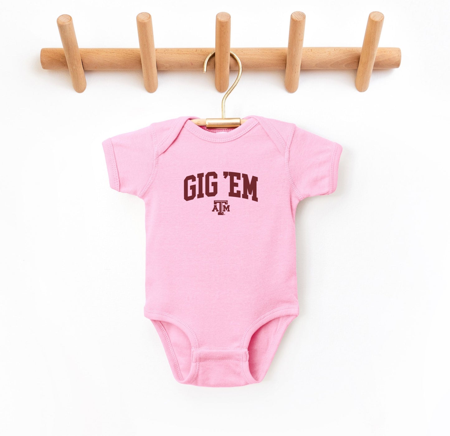 The Pink Infant Unisex Texas A&M Gig 'Em Collegiate Bodysuit lays flat on a white background. The ﻿Texas A&M Gig 'Em Collegiate﻿ graphic is in bold Maroon in a Collegiate style.