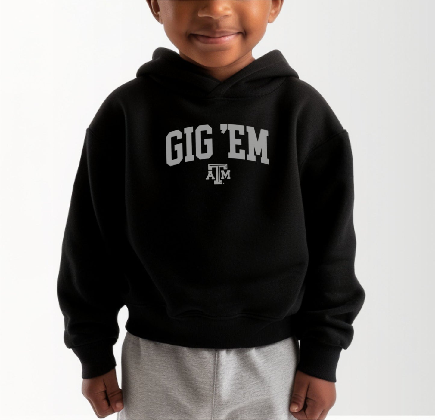 A model wears the Black Toddler Unisex Texas A&M Gig 'Em Collegiate Hooded Sweatshirt.  The ﻿Texas A&M Gig 'Em Collegiate﻿ graphic is in bold White in a Collegiate style.