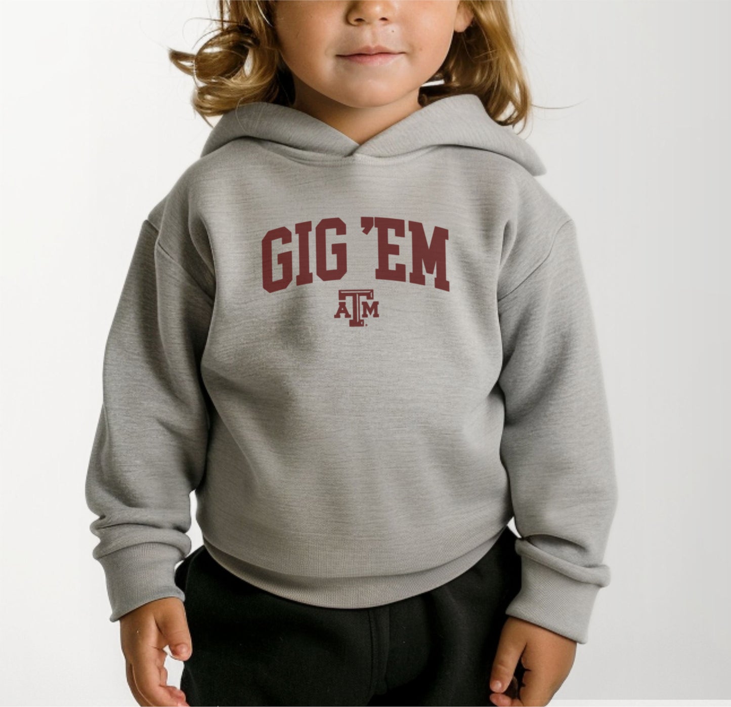 A model wears the Heather Grey Toddler Unisex Texas A&M Gig 'Em Collegiate Hooded Sweatshirt.  The ﻿Texas A&M Gig 'Em Collegiate﻿ graphic is in bold Maroon in a Collegiate style.