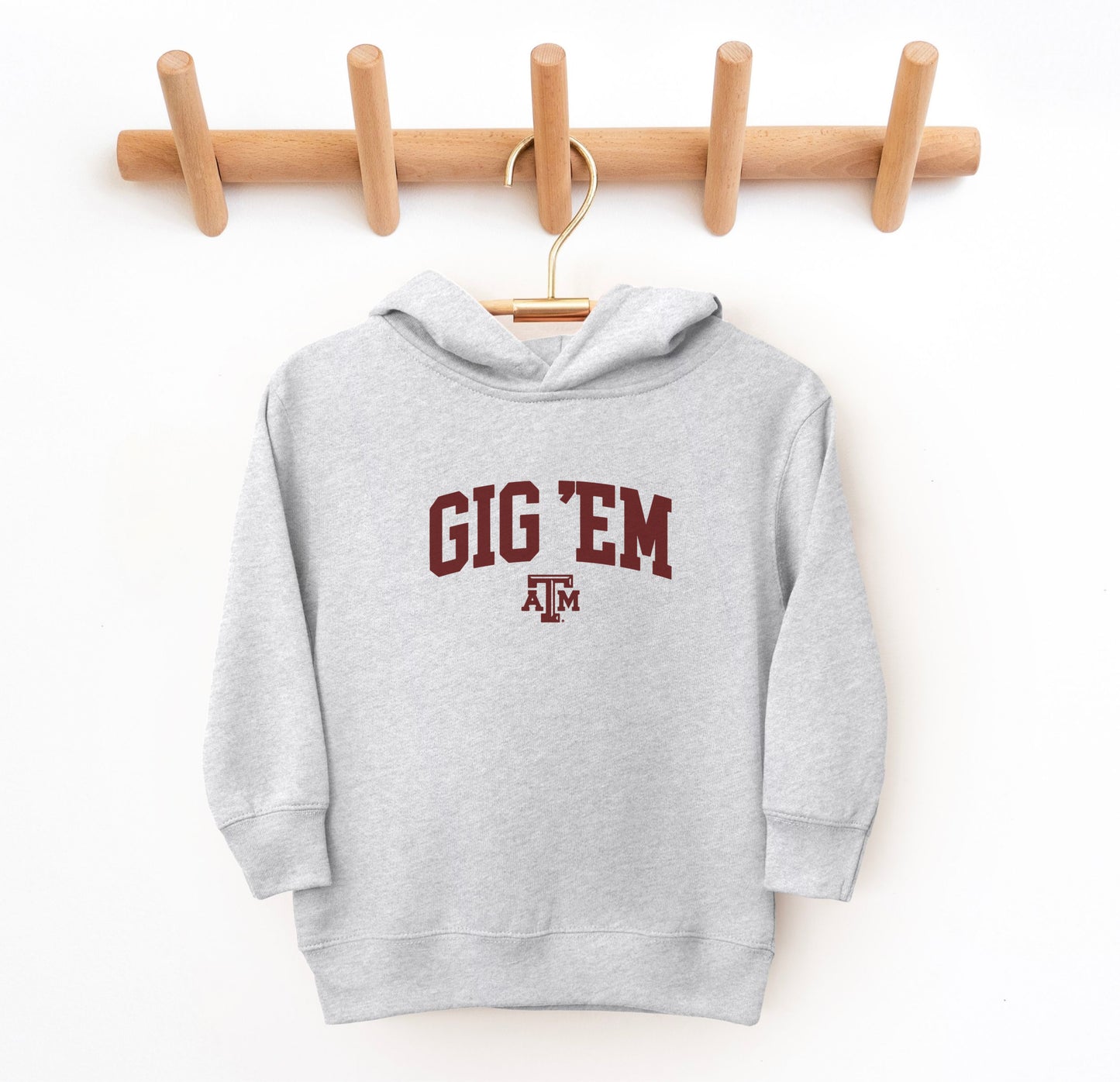 The Heather Grey Toddler Unisex Texas A&M Gig 'Em Collegiate Hooded Sweatshirt lays flat on a white background. The ﻿Texas A&M Gig 'Em Collegiate﻿ graphic is in bold Maroon in a Collegiate style.