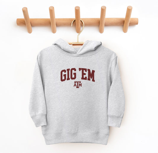 The Heather Grey Toddler Unisex Texas A&M Gig 'Em Collegiate Hooded Sweatshirt lays flat on a white background. The ﻿Texas A&M Gig 'Em Collegiate﻿ graphic is in bold Maroon in a Collegiate style.