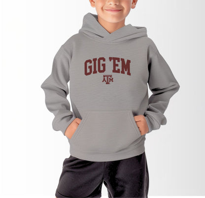 A model wears the Sport Grey Youth Unisex Texas A&M Gig 'Em Collegiate Hooded Sweatshirt.  The ﻿Texas A&M Gig 'Em Collegiate﻿ graphic is in bold Maroon in a Collegiate style.