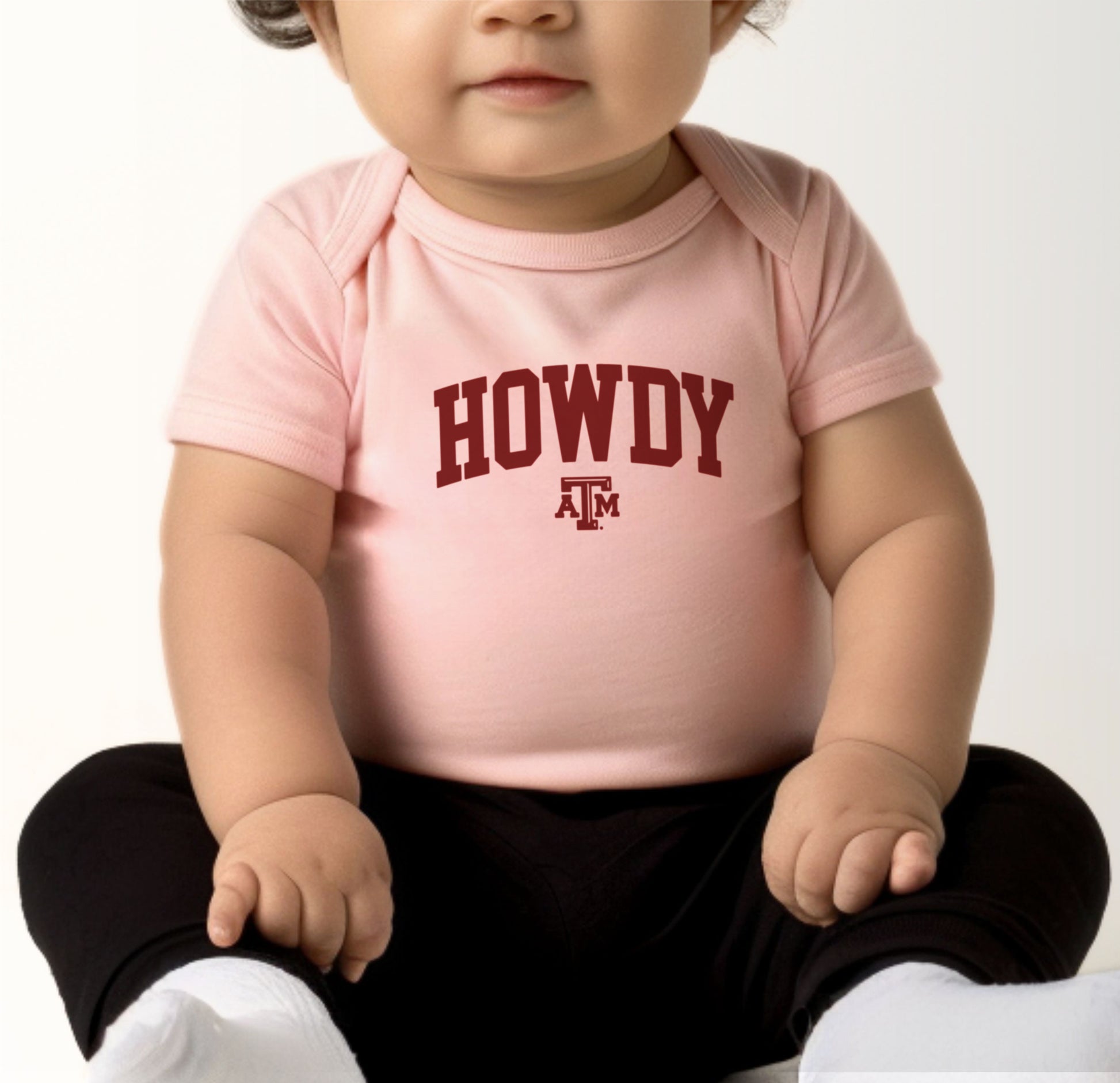 A model wears the Heather Grey Infant Unisex Texas A&M Howdy Varsity Bodysuit.  The ﻿Texas A&M Howdy Varsity﻿ graphic is in bold Maroon in a Collegiate style.