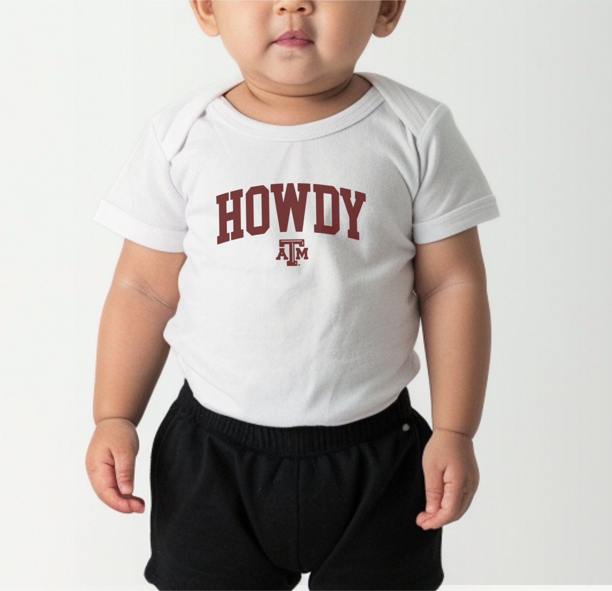 A model wears the Black Infant Unisex Texas A&M Howdy Varsity Bodysuit.  The ﻿Texas A&M Howdy Varsity﻿ graphic is in bold White in a Collegiate style.