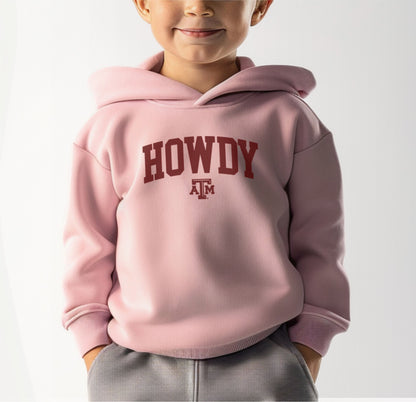 A model wears the Pink Toddler Unisex Texas A&M Howdy Varsity Hooded Sweatshirt.  The ﻿Texas A&M Howdy Varsity﻿ graphic is in bold White in a Collegiate style.