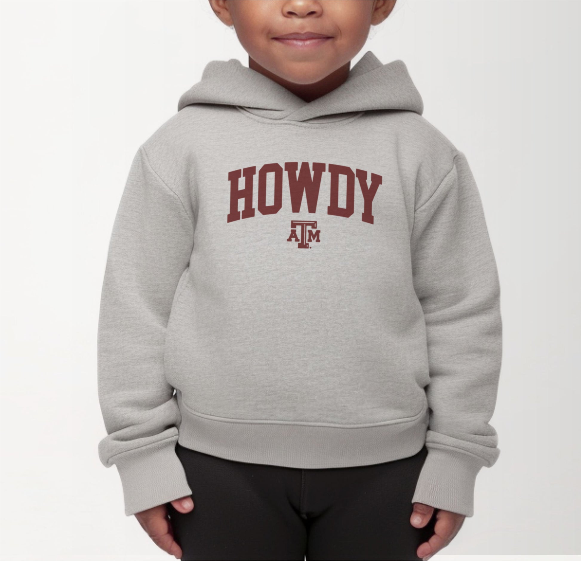 A model wears the Heather Grey Toddler Unisex Texas A&M Howdy Varsity Hooded Sweatshirt.  The ﻿Texas A&M Howdy Varsity﻿ graphic is in bold Maroon in a Collegiate style.