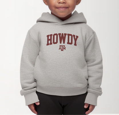 A model wears the Heather Grey Toddler Unisex Texas A&M Howdy Varsity Hooded Sweatshirt.  The ﻿Texas A&M Howdy Varsity﻿ graphic is in bold Maroon in a Collegiate style.