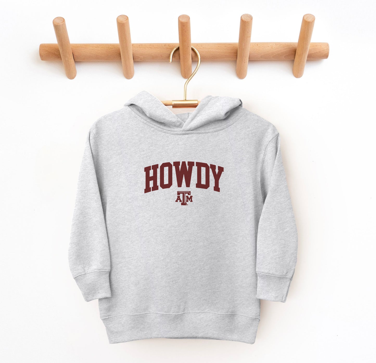 The Heather Grey Toddler Unisex Texas A&M Howdy Varsity Hooded Sweatshirt lays flat on a white background. The ﻿Texas A&M Howdy Varsity﻿ graphic is in bold Maroon in a Collegiate style.