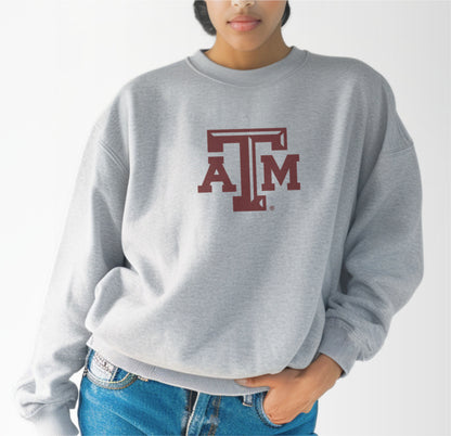 A model wears the Sport Grey Adult Unisex Texas A&M Logo Crewneck Sweatshirt.  The ﻿Texas A&M Logo﻿ graphic is in bold Maroon in a Varsity style.