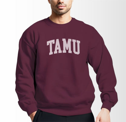 A model wears the Maroon Adult Unisex Texas A&M TAMU Crewneck Sweatshirt.  The ﻿Texas A&M TAMU﻿ graphic is in bold White in a Collegiate style.