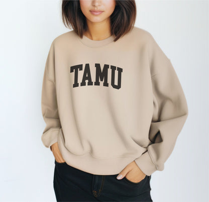 A model wears the Sand Adult Unisex Texas A&M TAMU Crewneck Sweatshirt.  The ﻿Texas A&M TAMU﻿ graphic is in bold Black in a Collegiate style.