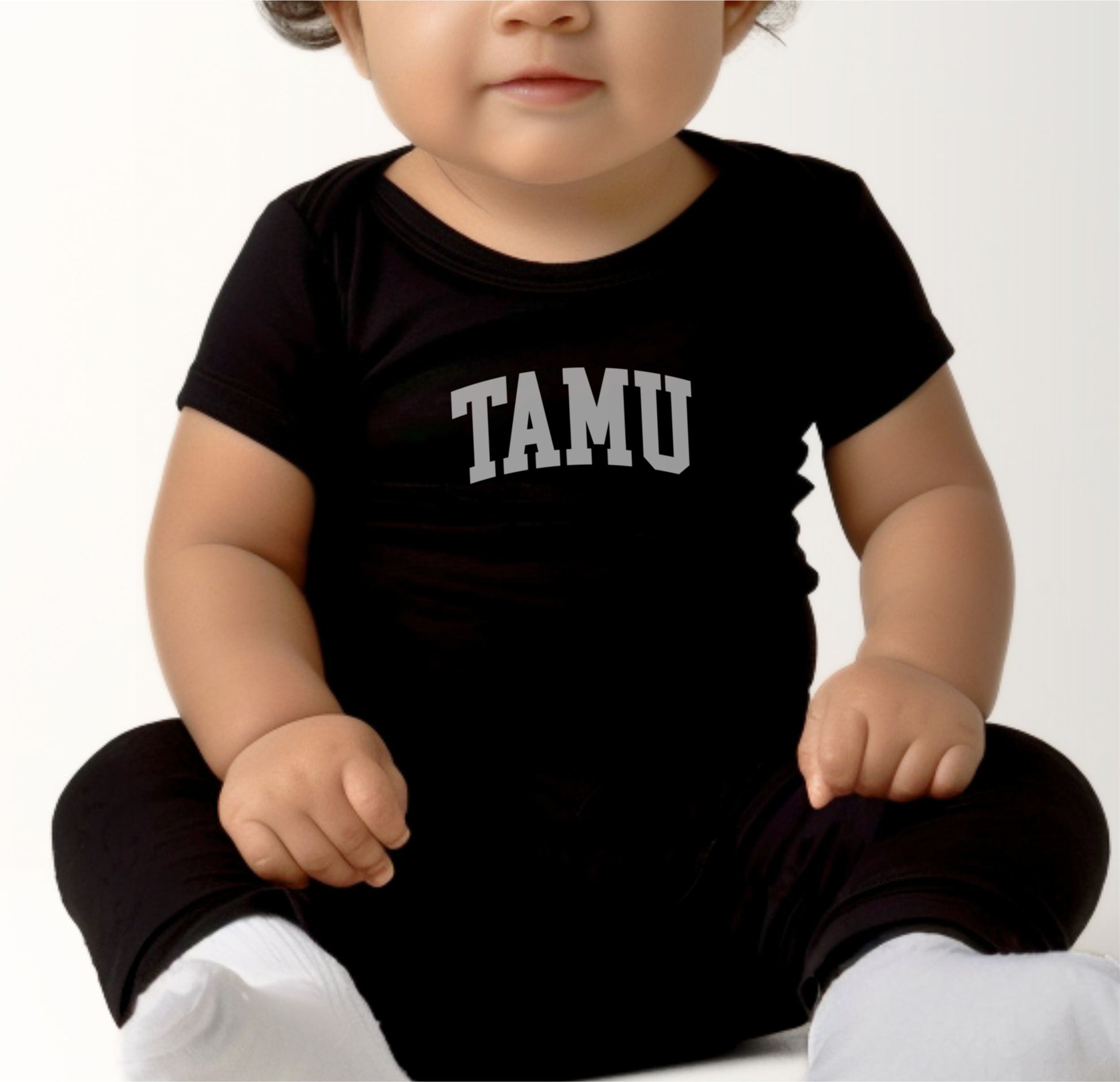 A model wears the Black Infant Unisex Texas A&M TAMU Bodysuit.  The ﻿Texas A&M TAMU﻿ graphic is in bold White in a Collegiate style.