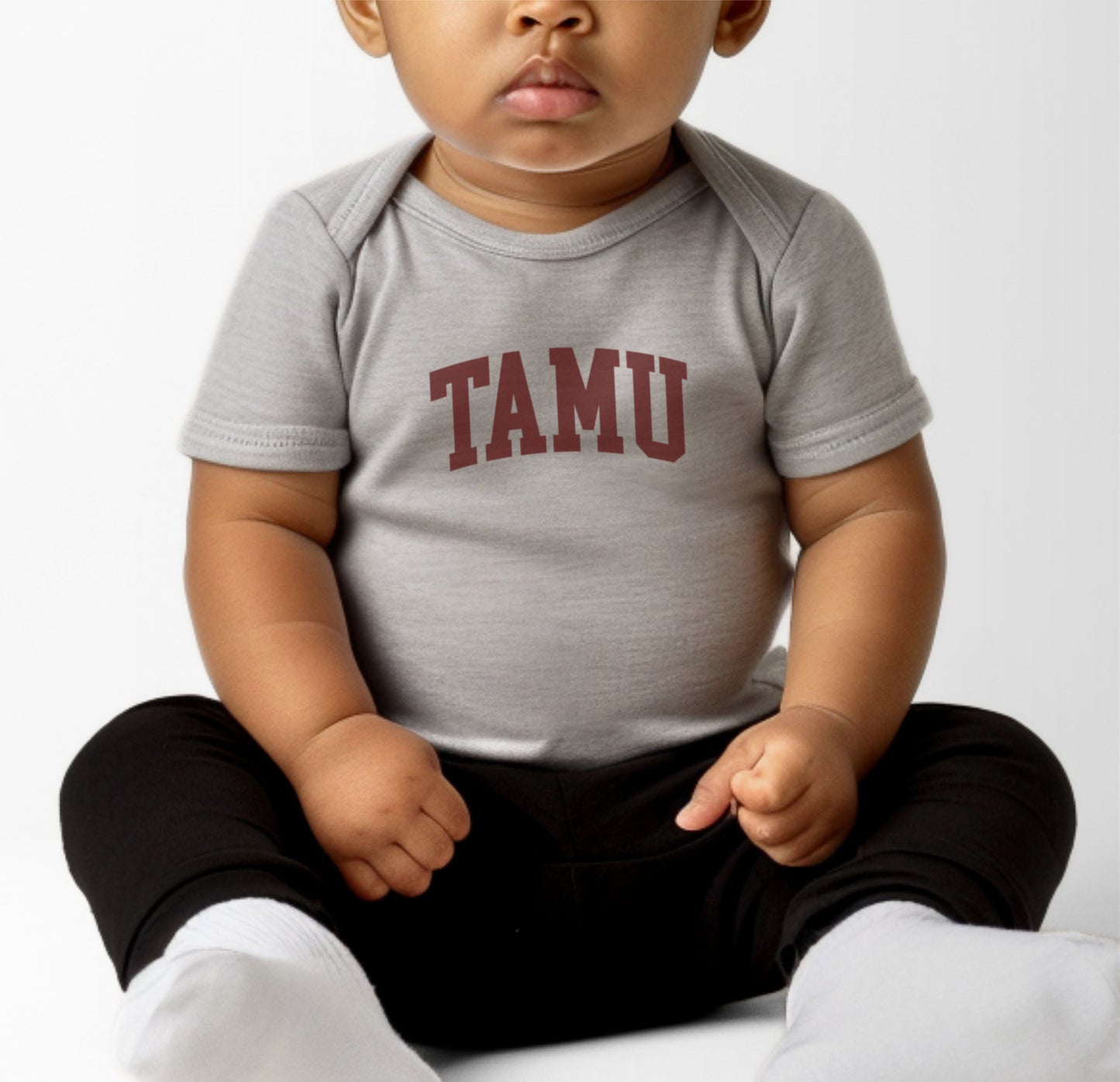 A model wears the Heather Grey Infant Unisex Texas A&M TAMU Bodysuit.  The ﻿Texas A&M TAMU﻿ graphic is in bold Maroon in a Collegiate style.