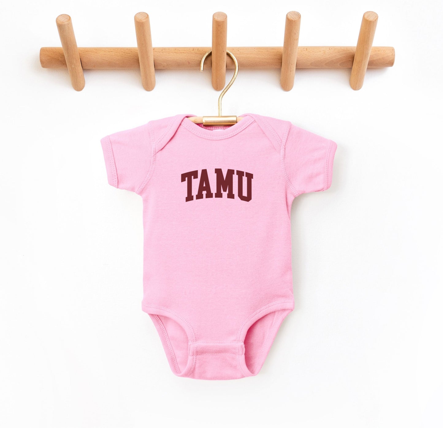 The Pink Infant Unisex Texas A&M TAMU Bodysuit lays flat on a white background. The ﻿Texas A&M TAMU﻿ graphic is in bold Maroon in a Collegiate style.