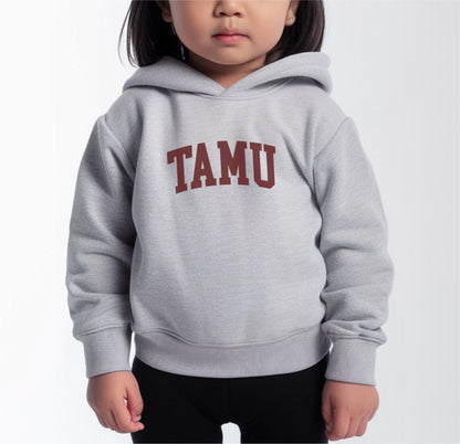 A model wears the Heather Grey Toddler Unisex Texas A&M TAMU Hooded Sweatshirt.  The ﻿Texas A&M TAMU﻿ graphic is in bold Maroon in a Collegiate style.