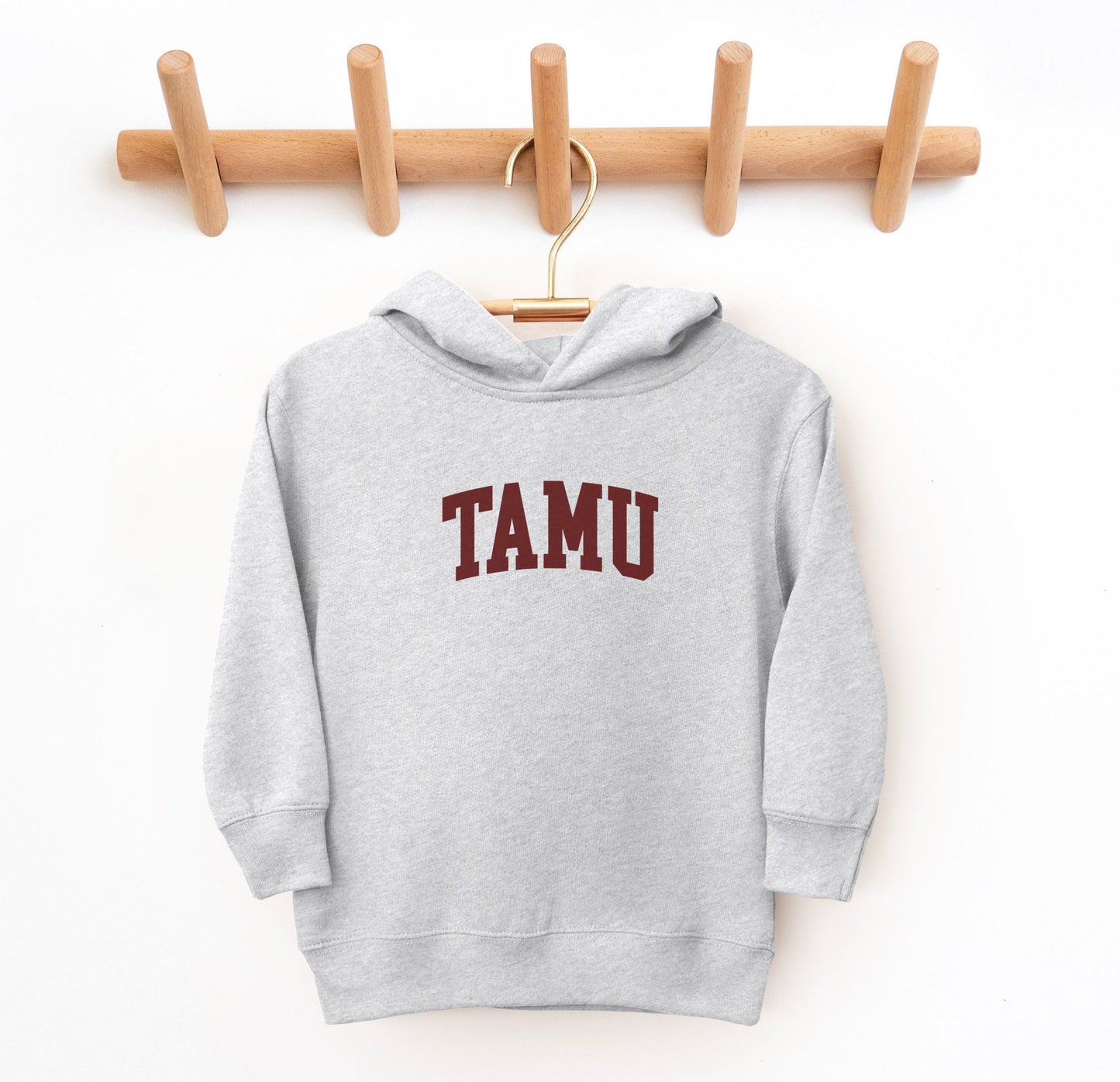 The Heather Grey Toddler Unisex Texas A&M TAMU Hooded Sweatshirt lays flat on a white background. The ﻿Texas A&M TAMU﻿ graphic is in bold Maroon in a Collegiate style.