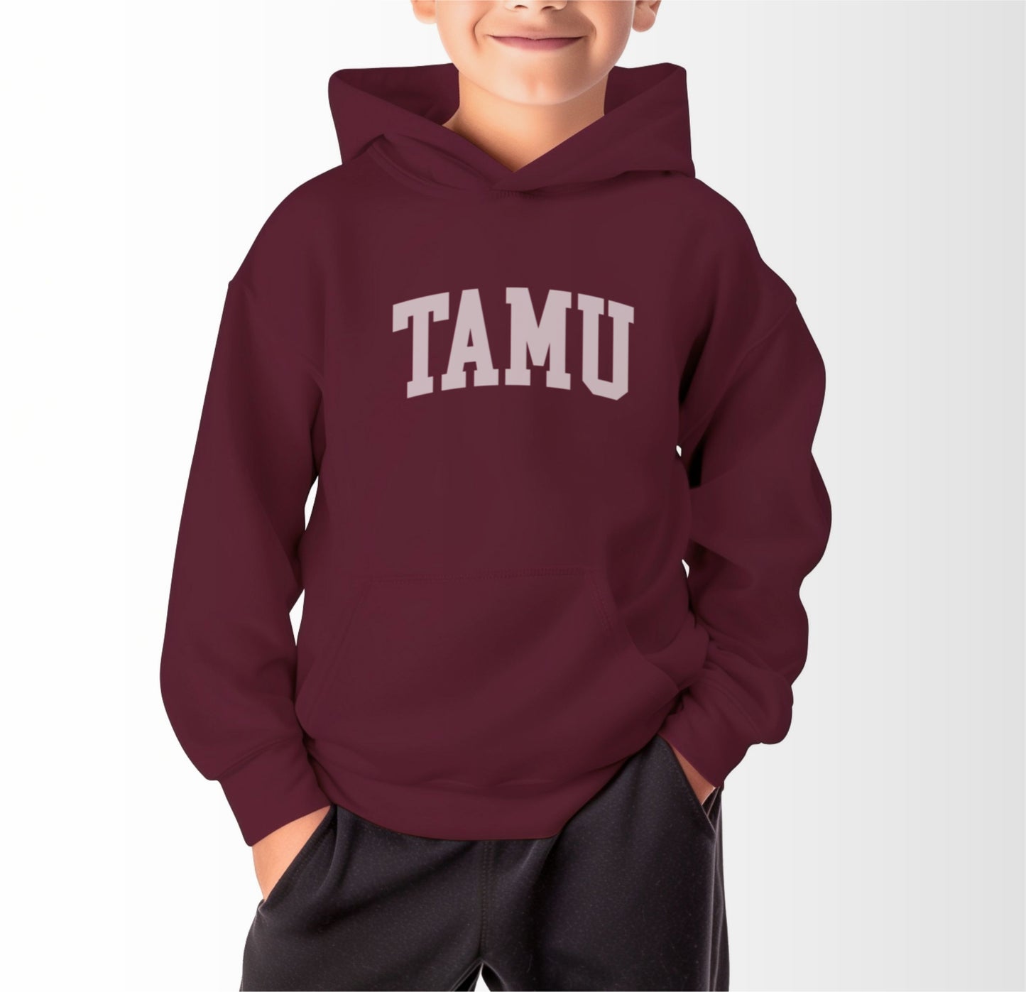 A model wears the Maroon Youth Unisex Texas A&M TAMU Hooded Sweatshirt.  The ﻿Texas A&M TAMU﻿ graphic is in bold White in a Collegiate style.