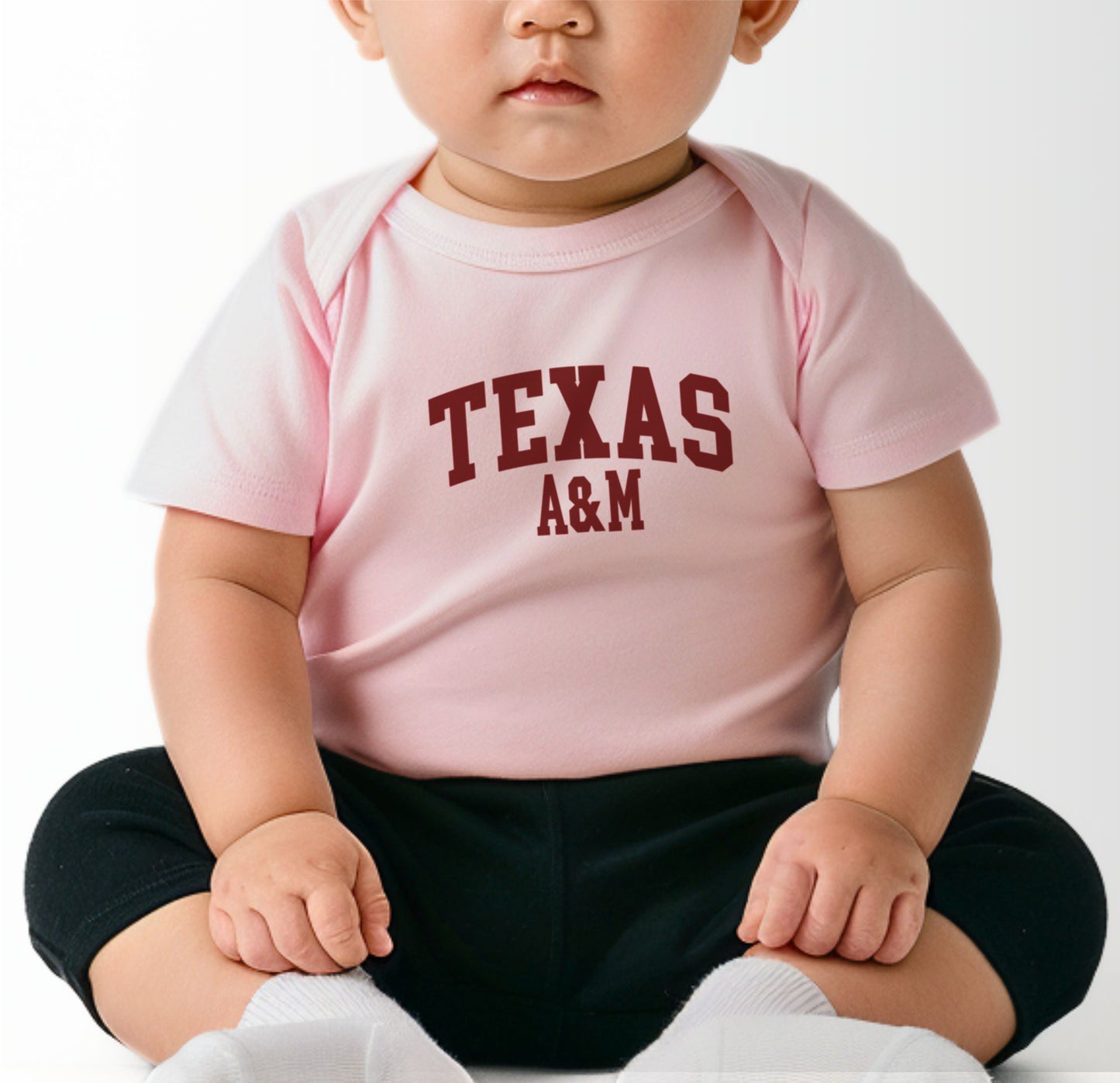 A model wears the Heather Grey Infant Unisex Texas A&M Varsity Bodysuit.  The ﻿Texas A&M Varsity﻿ graphic is in bold Maroon in a Collegiate style.