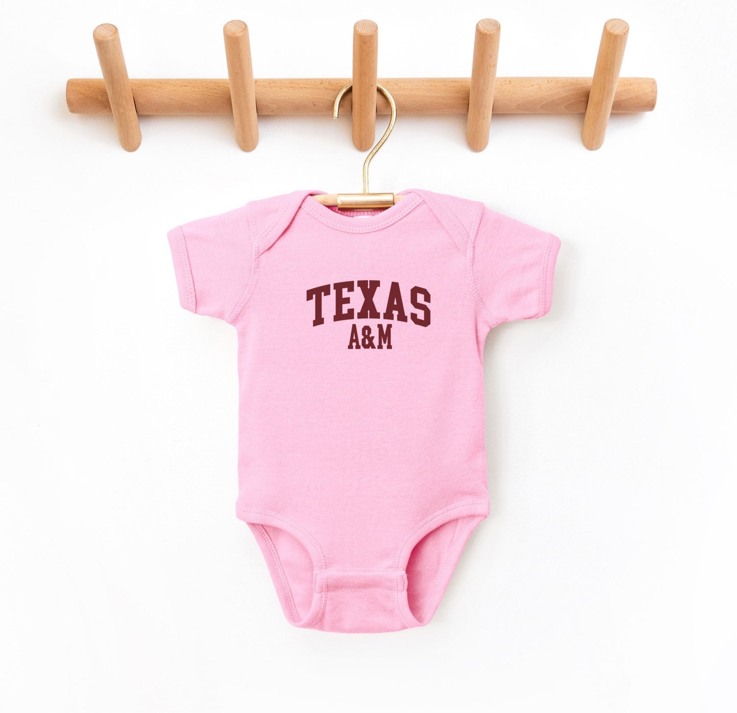 The Pink Infant Unisex Texas A&M Varsity Bodysuit lays flat on a white background. The ﻿Texas A&M Varsity﻿ graphic is in bold Maroon in a Collegiate style.