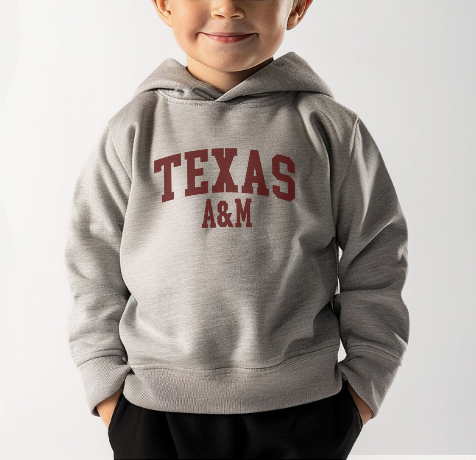 A model wears the Heather Grey Toddler Unisex Texas A&M Varsity Hooded Sweatshirt.  The ﻿Texas A&M Varsity﻿ graphic is in bold Maroon in a Collegiate style.