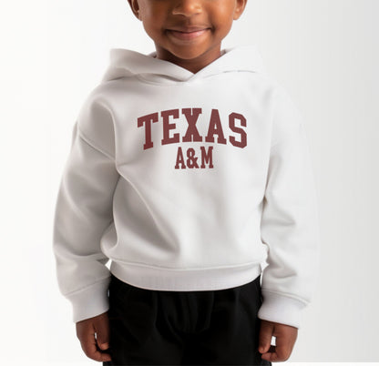 A model wears the White Toddler Unisex Texas A&M Varsity Hooded Sweatshirt.  The ﻿Texas A&M Varsity﻿ graphic is in bold Maroon in a Collegiate style.