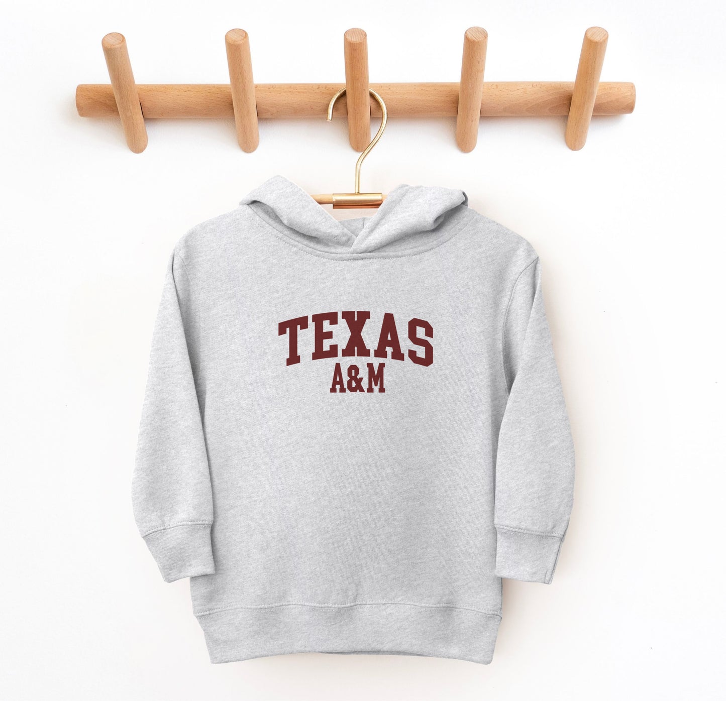 The Heather Grey Toddler Unisex Texas A&M Varsity Hooded Sweatshirt lays flat on a white background. The ﻿Texas A&M Varsity﻿ graphic is in bold Maroon in a Collegiate style.