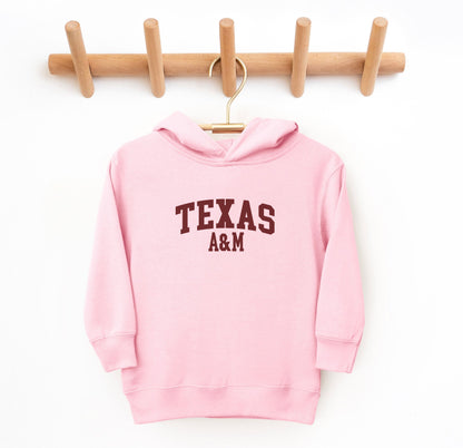 The Pink Toddler Unisex Texas A&M Varsity Hooded Sweatshirt lays flat on a white background. The ﻿Texas A&M Varsity﻿ graphic is in bold White in a Collegiate style.