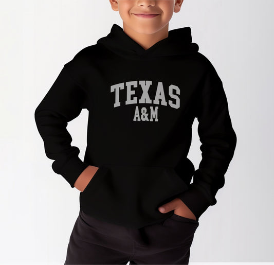 A model wears the Black Youth Unisex Texas A&M Varsity Hooded Sweatshirt.  The ﻿Texas A&M Varsity﻿ graphic is in bold White in a Collegiate style.
