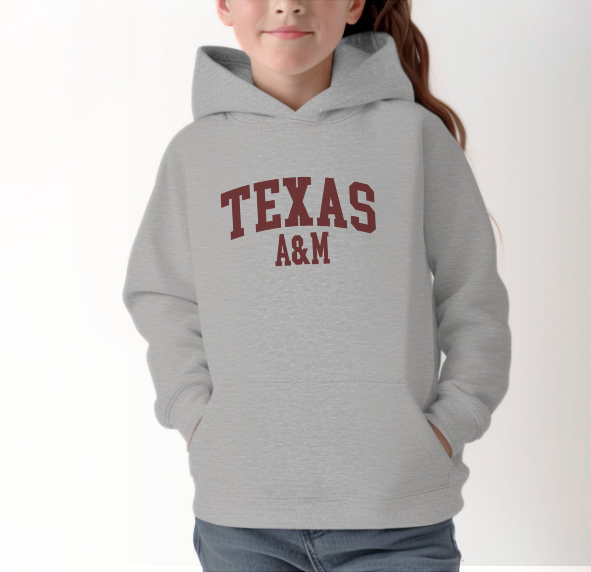 A model wears the Sport Grey Youth Unisex Texas A&M Varsity Hooded Sweatshirt.  The ﻿Texas A&M Varsity﻿ graphic is in bold Maroon in a Collegiate style.