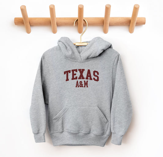 The Grey Youth Unisex Texas A&M Varsity Hooded Sweatshirt lays flat on a white background. The ﻿Texas A&M Varsity﻿ graphic is in bold Maroon in a Collegiate style.