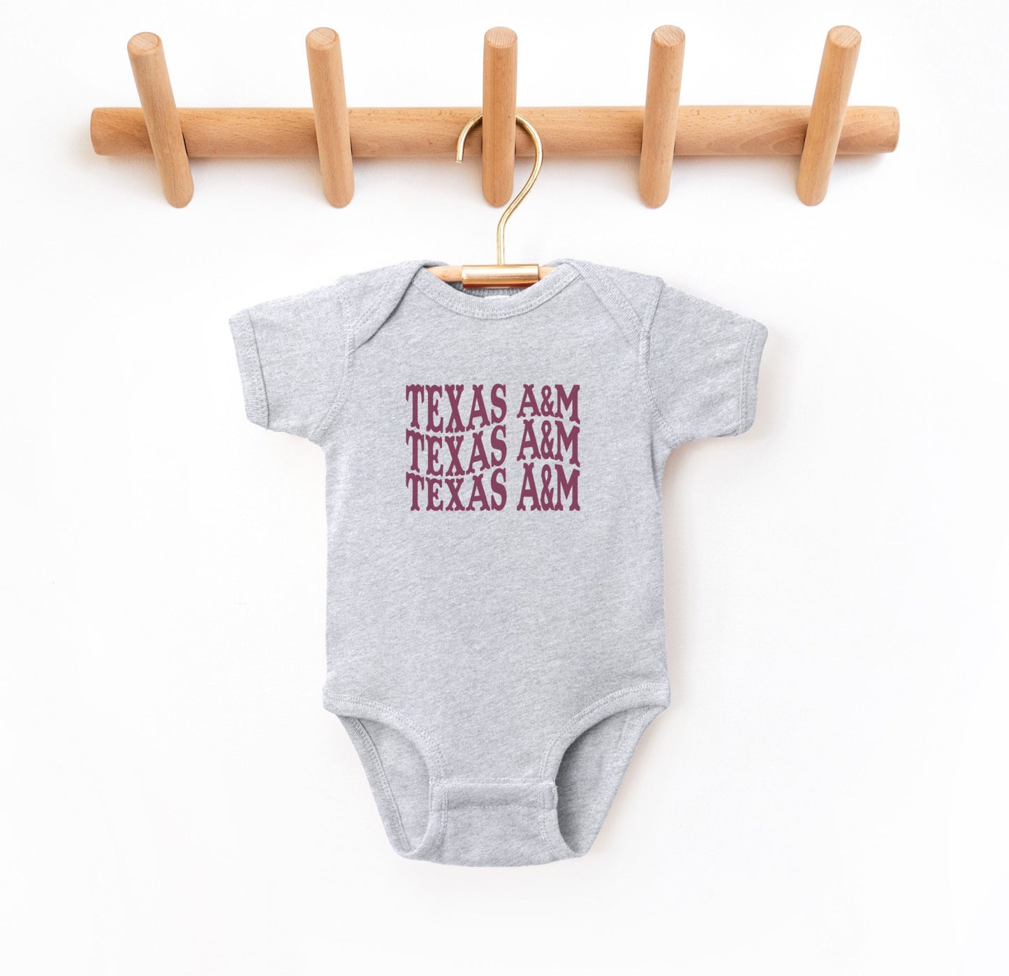 The Heather Grey Infant Unisex Texas A&M Western Bodysuit lays flat on a white background. The ﻿Texas A&M Western﻿ graphic is in bold Maroon in a Western style.