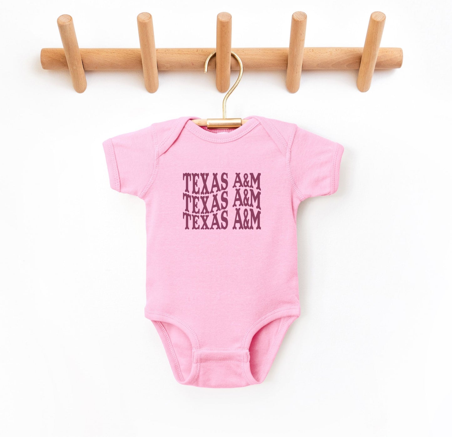 The Pink Infant Unisex Texas A&M Western Bodysuit lays flat on a white background. The ﻿Texas A&M Western﻿ graphic is in bold Maroon in a Western style.