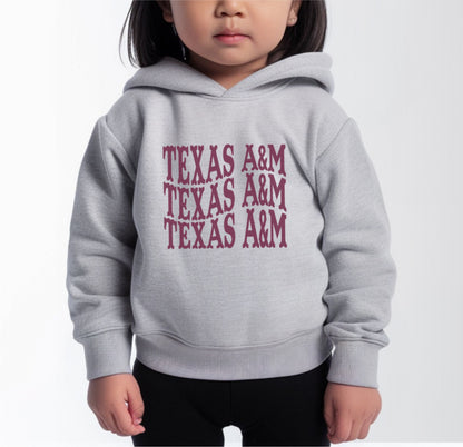 A model wears the Heather Grey Toddler Unisex Texas A&M Western Hooded Sweatshirt.  The ﻿Texas A&M Western﻿ graphic is in bold Maroon in a Western style.