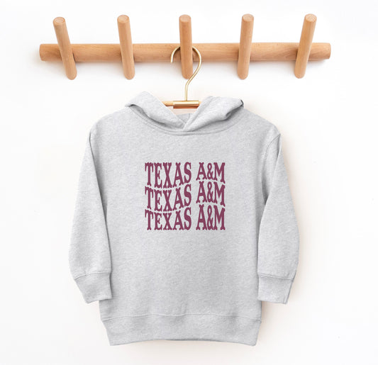 The Heather Grey Toddler Unisex Texas A&M Western Hooded Sweatshirt lays flat on a white background. The ﻿Texas A&M Western﻿ graphic is in bold Maroon in a Western style.