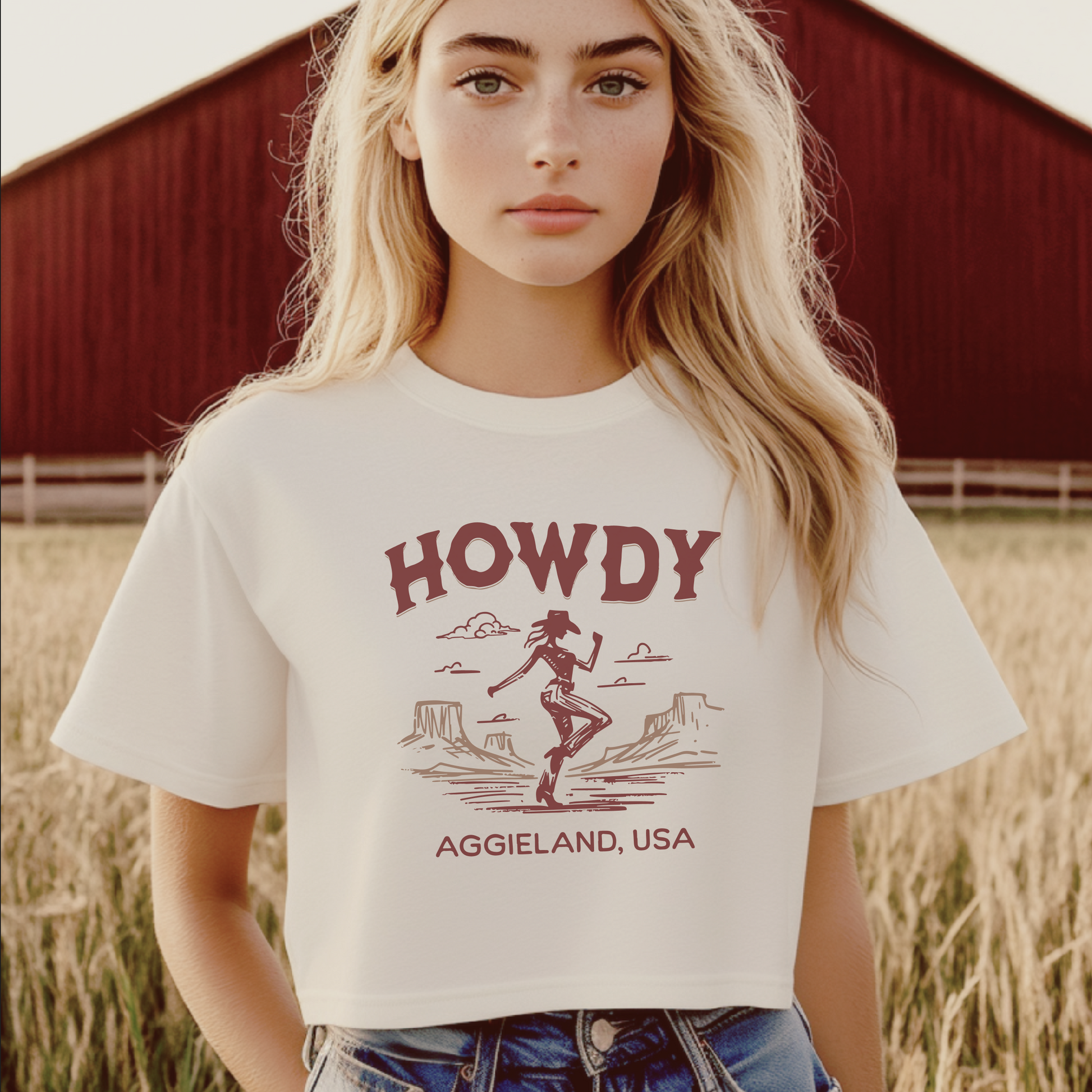 A white cropped t-shirt with the word "HOWDY" in maroon letters at the top. Below, a maroon silhouette of a cowgirl dancing in front of a desert landscape with mountains. The text "AGGIE LAND, USA" is written in maroon at the bottom. The model stands in a field on a Texas Ranch. 