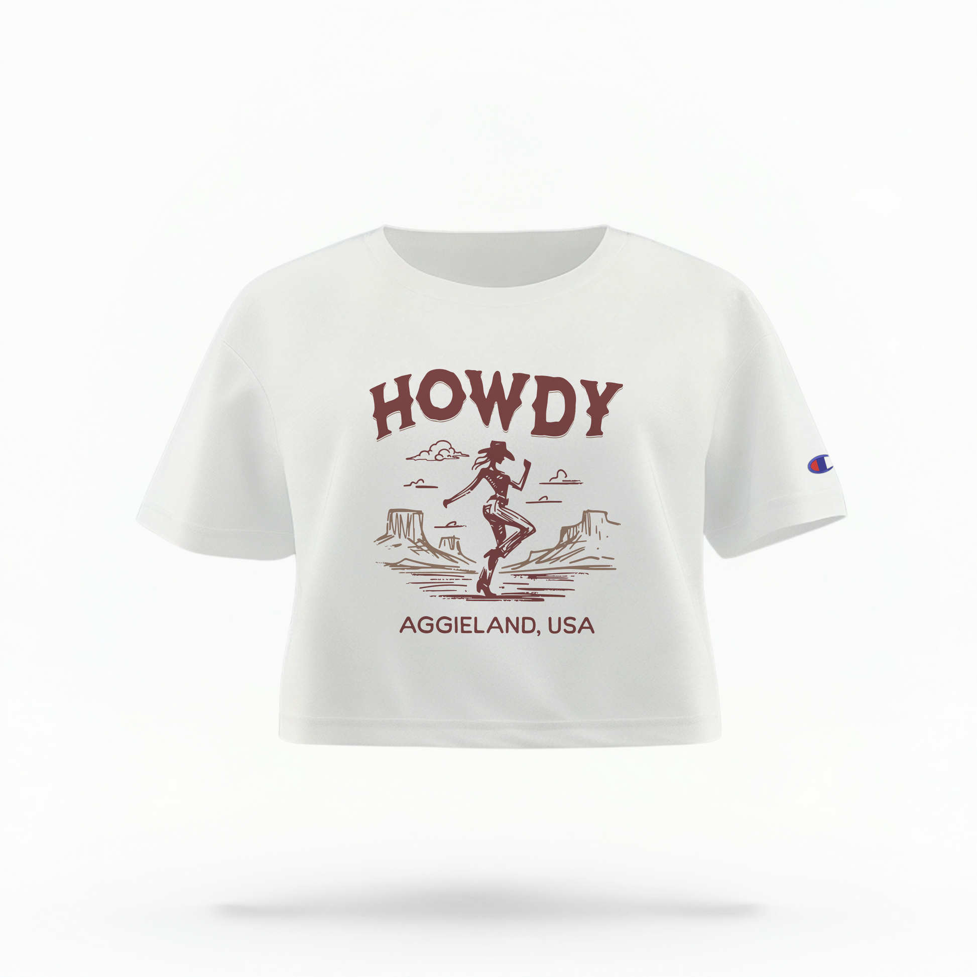 A white cropped t-shirt with the word "HOWDY" in maroon letters at the top. Below, a maroon silhouette of a cowgirl in front of a desert landscape with mountains. The text "AGGIE LAND, USA" is written in maroon at the bottom. The shirt is a Champion brand.