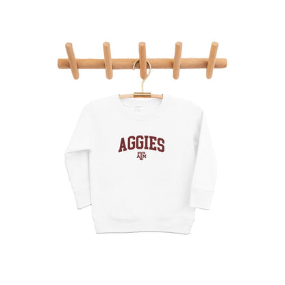 The white Toddler Texas A&M Aggies Collegiate Crewneck Sweatshirt hangs from a pine clothing rack.  The ﻿Texas A&M Aggies Collegiate﻿ graphic is in bold Maroon in a Varsity style.