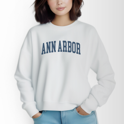 A model wears the White Adult Unisex Ann Arbor Michigan Varsity Crewneck Sweatshirt. The Ann Arbor Michigan Varsity graphic is in bold Navy in a Collegiate style.