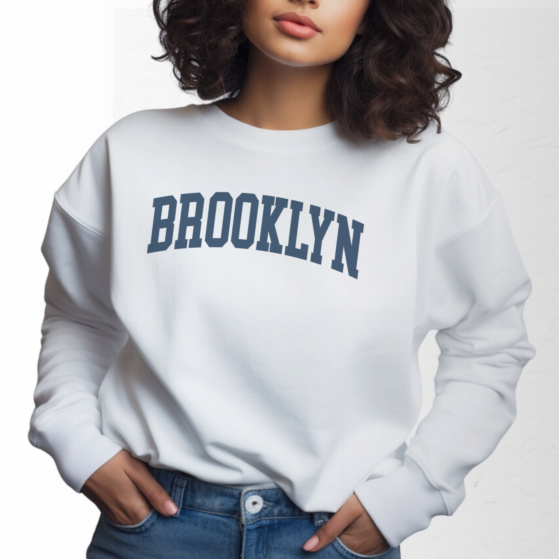 A model wears the White Adult Unisex Brooklyn NY Collegiate Crewneck Sweatshirt.  The ﻿Brooklyn NY Collegiate﻿ graphic is in bold Navy in a Varsity style.