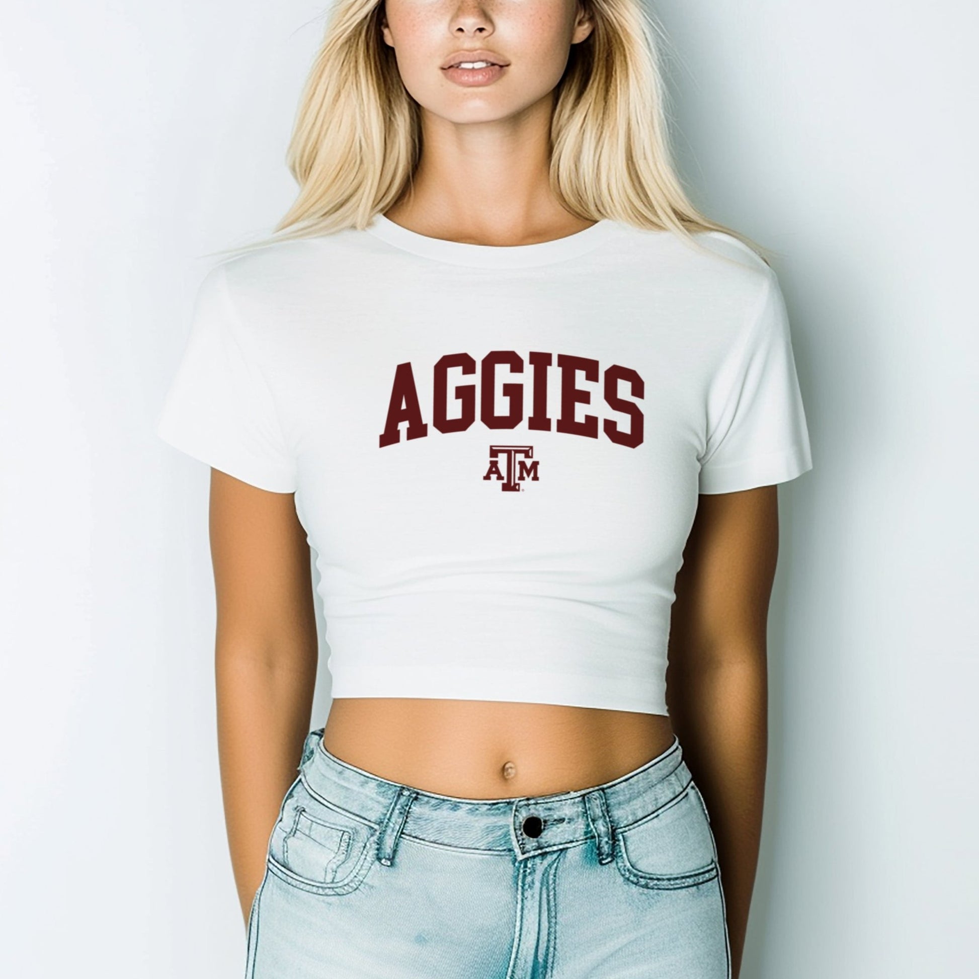 A model wears the White Texas A&M Aggies Collegiate Crop Top.  The ﻿Texas A&M Aggies Collegiate﻿ graphic is in bold Maroon in a Varsity style.