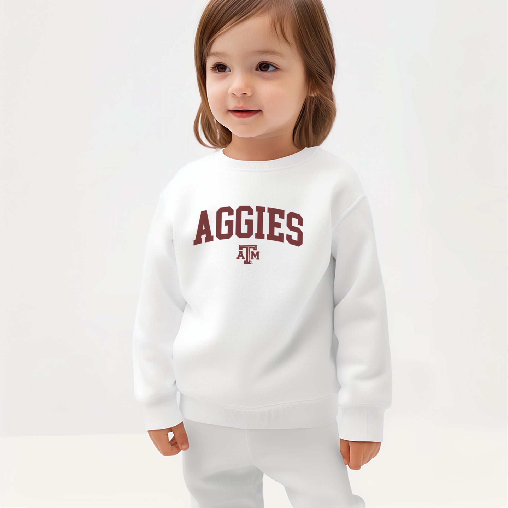 A young toddler wears the White Texas A&M Aggies Collegiate Crewneck Sweatshirt.  The ﻿Texas A&M Aggies Collegiate﻿ graphic is in bold Maroon in a Varsity style.